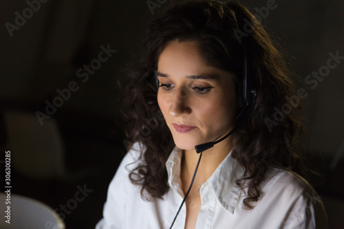 Focused young businesswoman in headset. Close-up view of concentrated female call center operator working late in dark office. Client service concept