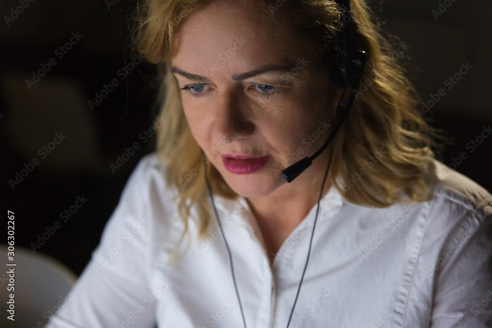 Serious businesswoman in headset looking down. Professional female call center operator in headset working late in dark office. Customer service concept Serious businesswoman in headset looking down. Professional female call center operator in headset working late in dark office. Customer service concept