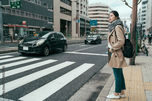 pretty asian chinese woman traveler walking in city osaka waiting for traffic lights to cross busy road with cars driving by. full length girl with bag standing on street and ready to pass pedestrian photo