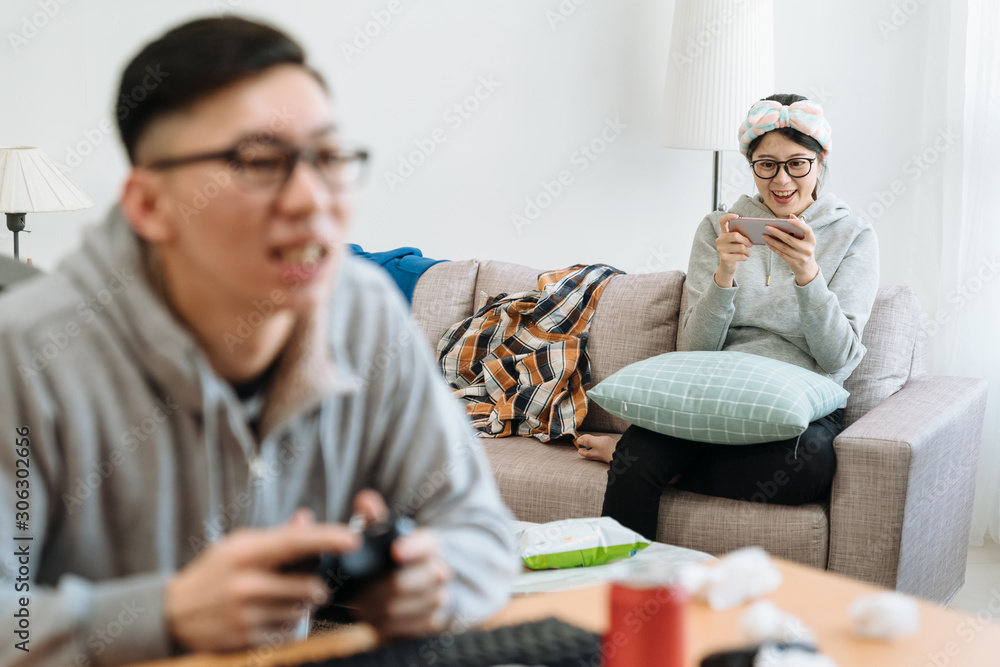 Happy couple having fun in themselves at home. bokeh view of young man playing video online games on computer with joystick. smiling cute girl friend laughing watching comedy on smart phone on couch Happy couple having fun in themselves at home. bokeh view of young man playing video online games on computer with joystick. smiling cute girl friend laughing watching comedy on smart phone on couch