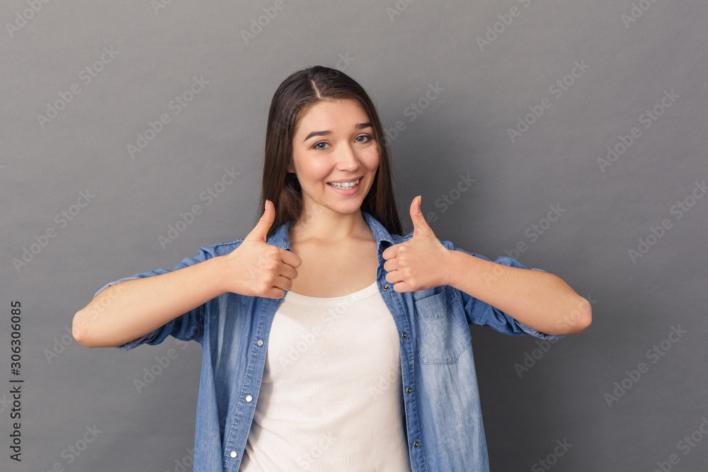 Freestyle. Young woman in denim shirt standing studio isolated on grey thumbs up smiling cheerful