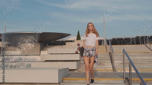 A young blonde girl, a model with a longboard in her hand in a white shirt and denim shorts goes down the stairs and poses for the camera. Urban modern structure in the background.