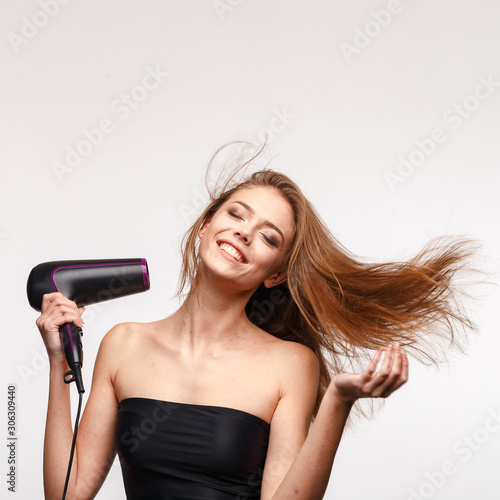 A young attractive girl is blow-drying her hair and laughing.