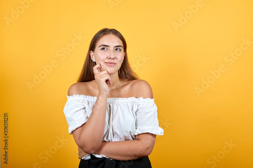 Attractive young womankeeping hand on chin, thinking about issue isolated on orange background in studio in casual white shirt. People sincere emotions, lifestyle concept.