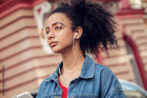 Outdoors Leisure. Young stylish african girl in denim jacket and glasses standing with earphones listening to music pensive bottom view close-up