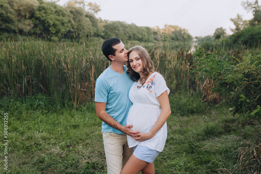 Portrait of a young family expecting a baby. Loving man and beautiful pregnant blond woman cuddling against the backdrop of greenery in the park. Pregnancy picture, concept.