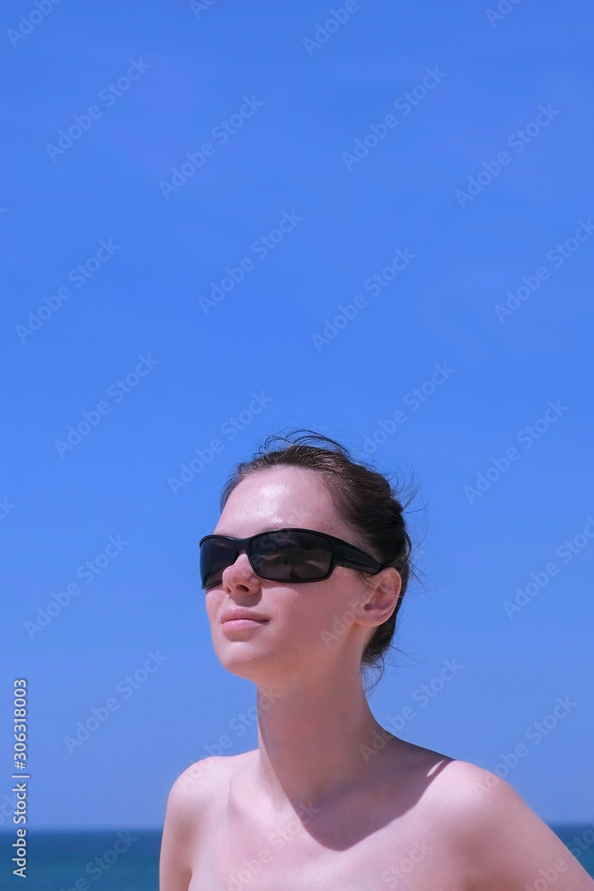 Portrait young woman sunbathing stand on beach on sea and blue sky background. Brunette girl is in sunglasses and bikini, front view. Turist travel summer vacation. Vertical Portrait young woman sunbathing stand on beach on sea and blue sky background. Brunette girl is in sunglasses and bikini, front view. Turist travel summer vacation. Vertical