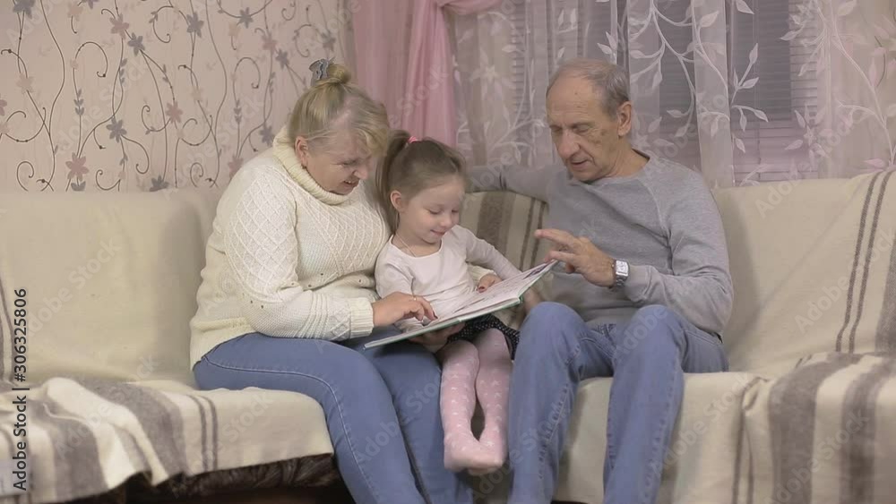 grandfather and grandmother are reading a book with their granddaughter sitting at home on the couch, family values grandfather and grandmother are reading a book with their granddaughter sitting at home on the couch, family values