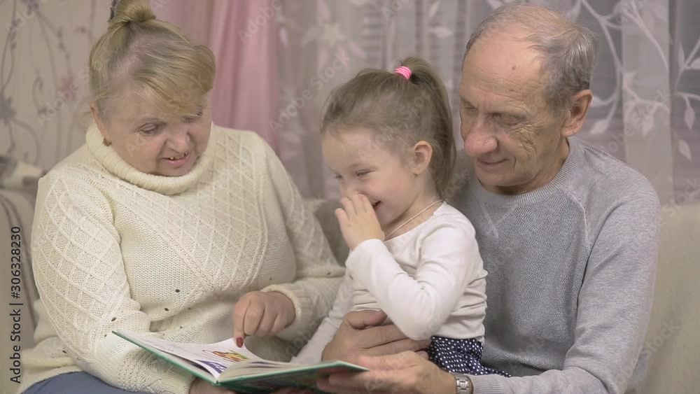 grandfather and grandmother are reading a book with their granddaughter sitting at home on the couch, family values grandfather and grandmother are reading a book with their granddaughter sitting at home on the couch, family values