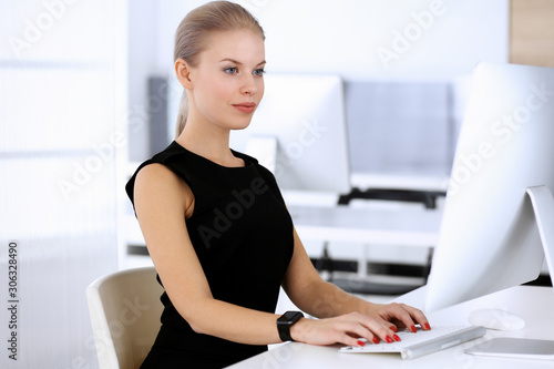 Business woman working with computer while sitting at the desk in modern office. Secretary or female lawyer looks beautiful in black dress. Business people concept