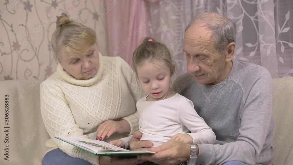 grandfather and grandmother are reading a book with their granddaughter sitting at home on the couch, family values grandfather and grandmother are reading a book with their granddaughter sitting at home on the couch, family values