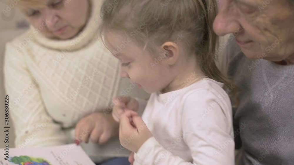 grandfather and grandmother are reading a book with their granddaughter sitting at home on the couch, family values grandfather and grandmother are reading a book with their granddaughter sitting at home on the couch, family values