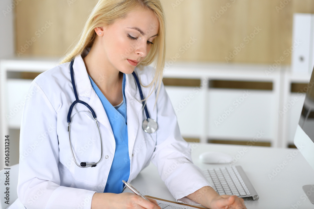 Doctor woman at work while sitting at the desk in hospital or clinic. Blonde cheerful physician filling up medical records form. Data and best service in medicine and healthcare Doctor woman at work while sitting at the desk in hospital or clinic. Blonde cheerful physician filling up medical records form. Data and best service in medicine and healthcare