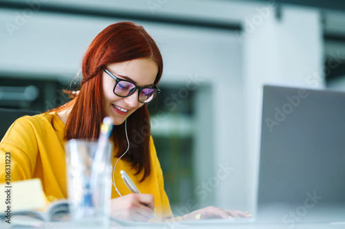 Young girl works at a computer in the office. Beautiful freelancer working from a laptop computer. Teenager student at the computer listens music to earphones and prepares for exams. Business woman.