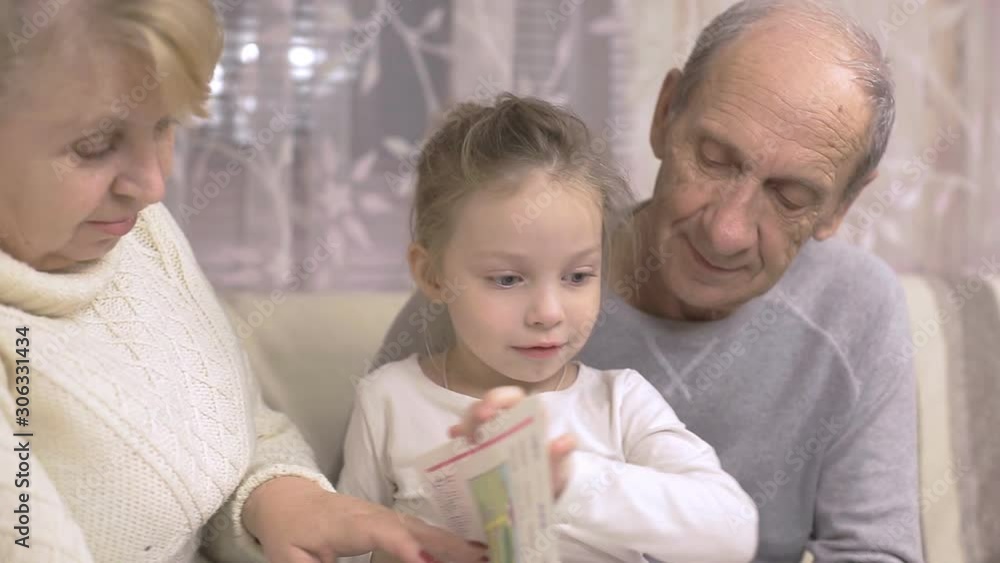 grandfather and grandmother are reading a book with their granddaughter sitting at home on the couch, family values grandfather and grandmother are reading a book with their granddaughter sitting at home on the couch, family values