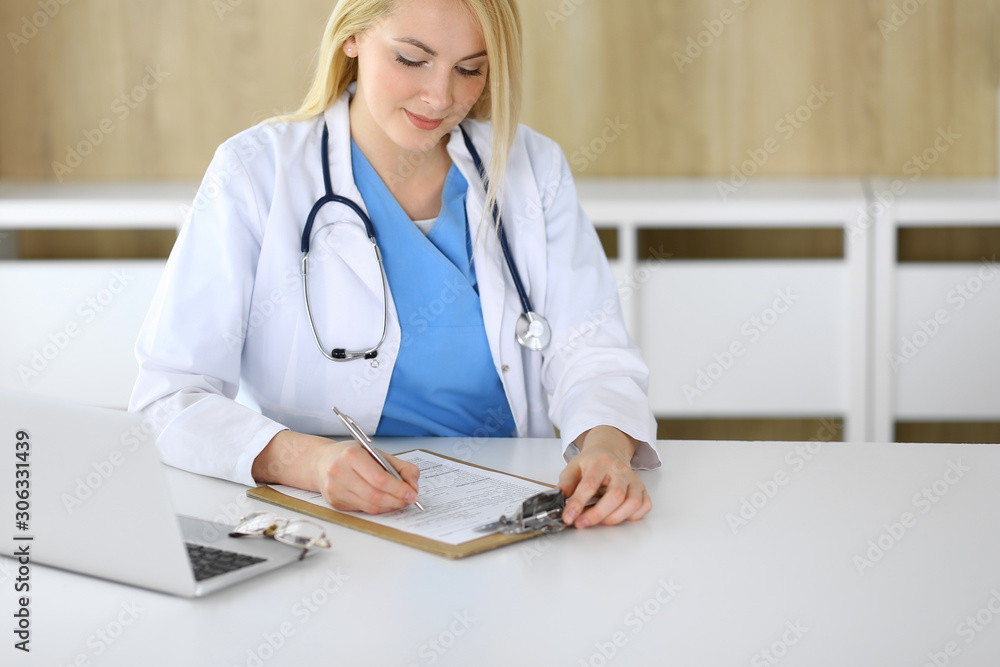Doctor woman at work while sitting at the desk in hospital or clinic. Blonde cheerful physician filling up medical records form. Data and best service in medicine and healthcare Doctor woman at work while sitting at the desk in hospital or clinic. Blonde cheerful physician filling up medical records form. Data and best service in medicine and healthcare