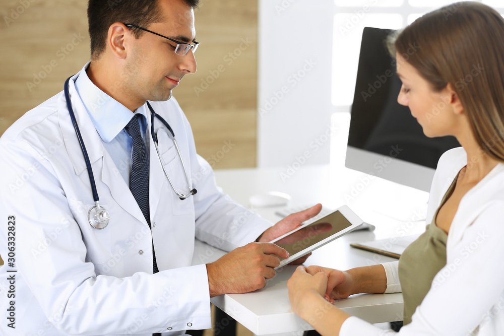 Doctor and patient discussing medical exam results while sitting at the desk in clinic. Male physician using tablet computer for filling up medication history records of young woman. Data in medicine Doctor and patient discussing medical exam results while sitting at the desk in clinic. Male physician using tablet computer for filling up medication history records of young woman. Data in medicine