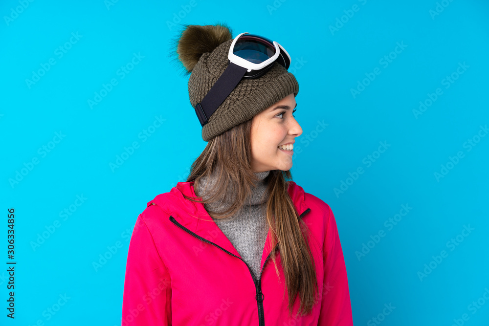 Teenager skier girl with snowboarding glasses over isolated blue background looking to the side