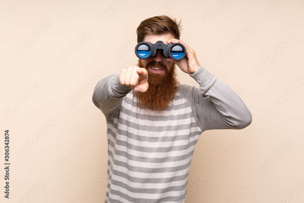 Redhead man with long beard over isolated background with black binoculars Redhead man with long beard over isolated background with black binoculars