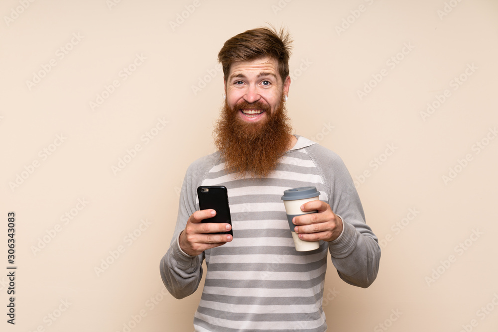 Redhead man with long beard over isolated background holding coffee to take away and a mobile Redhead man with long beard over isolated background holding coffee to take away and a mobile