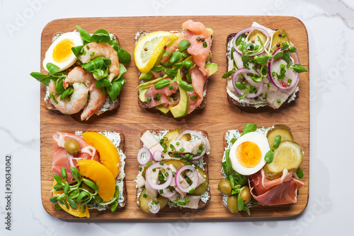 top view of wooden cutting board with tasty smorrebrod sandwiches on white marble surface