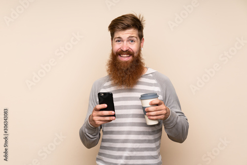 Redhead man with long beard over isolated background holding coffee to take away and a mobile