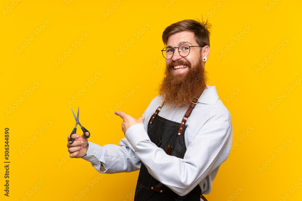 Barber with long beard in an apron over isolated yellow background and pointing it Barber with long beard in an apron over isolated yellow background and pointing it