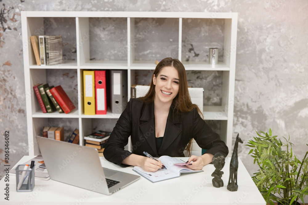 smiling beautiful woman manager with laptop sitting at her working place in office taking notes writing by hand in her planner looking happy. work concept smiling beautiful woman manager with laptop sitting at her working place in office taking notes writing by hand in her planner looking happy. work concept
