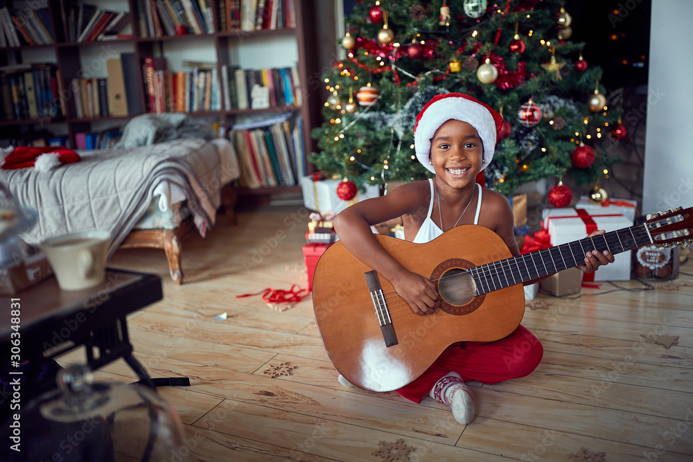 Fototapeta premium Girl in front of decoration Christmas tree play guitar.