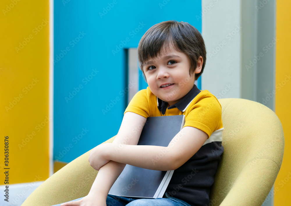 Portrait of Positive Child boy sitting on yellow chair holding his favorite book, A happy school kid with smilig face enjoying with reading time in the classroom. Education concept Portrait of Positive Child boy sitting on yellow chair holding his favorite book, A happy school kid with smilig face enjoying with reading time in the classroom. Education concept