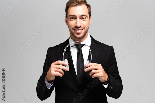 smiling businessman in suit holding headphones isolated on grey