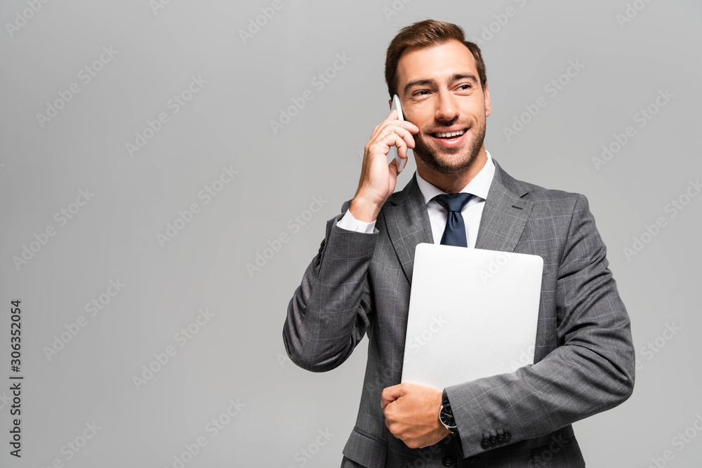 handsome and smiling businessman in suit holding laptop and talking on smartphone isolated on grey handsome and smiling businessman in suit holding laptop and talking on smartphone isolated on grey