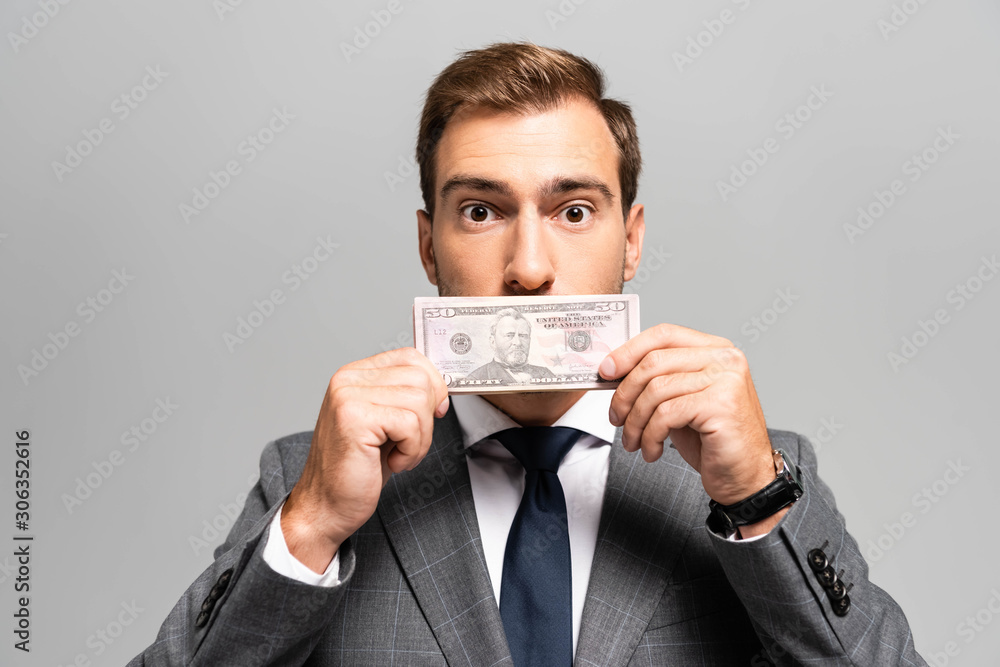 handsome businessman in suit obscuring face with dollar banknote isolated on grey
