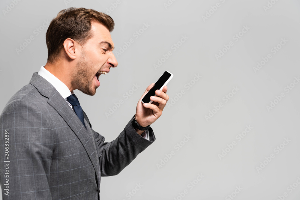 side view of handsome and angry businessman in suit shouting at smartphone isolated on grey side view of handsome and angry businessman in suit shouting at smartphone isolated on grey