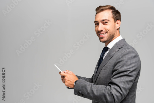 smiling and handsome businessman in suit using smartphone isolated on grey