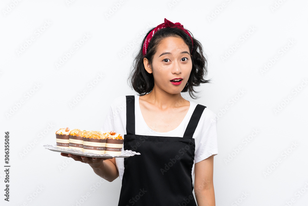 Young asian girl holding lots of muffin cake over isolated white background with surprise and shocked facial expression Young asian girl holding lots of muffin cake over isolated white background with surprise and shocked facial expression