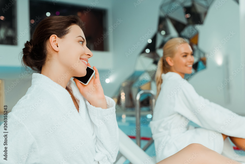 selective focus of attractive and smiling woman talking on smartphone and friend on background in spa selective focus of attractive and smiling woman talking on smartphone and friend on background in spa