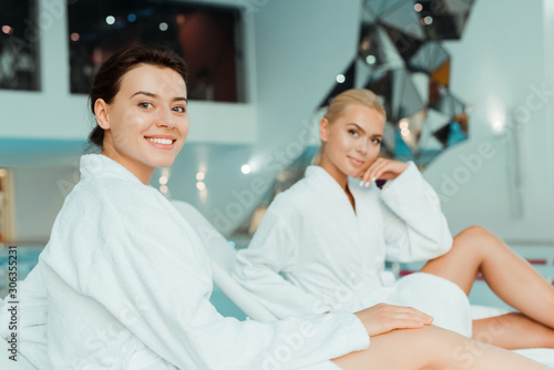 selective focus of attractive and smiling woman looking at camera and friend on background in spa