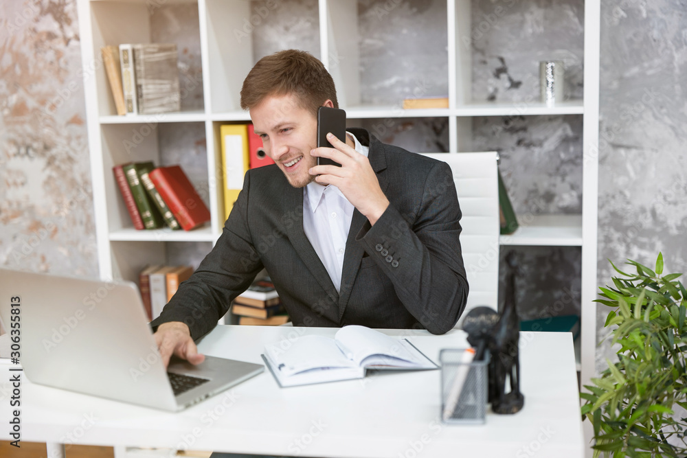 smiling handsome man manager checking mail in laptop and having phone conversation sitting at his working place in office looking involved. multitasking, work concept smiling handsome man manager checking mail in laptop and having phone conversation sitting at his working place in office looking involved. multitasking, work concept