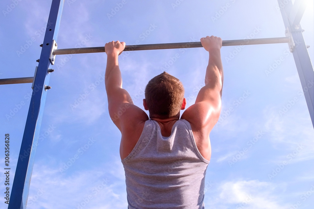 Fototapeta premium Sportsman bodybuilder making pull ups on horizontal bar in city park in sportsground on blue sky background, back view. Sport and healthy lifestyle. Independent fitness training and discipline.
