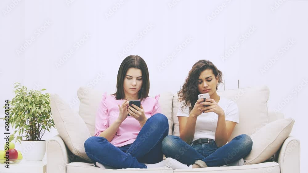 Beautiful young kinky afro-american and brunette caucasian girls are sitting on the sofa in lotos position, with phone in their hands, messaging. Beautiful young kinky afro-american and brunette caucasian girls are sitting on the sofa in lotos position, with phone in their hands, messaging.