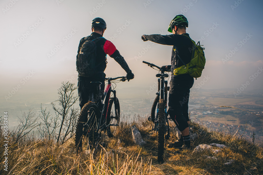 Naklejka premium Two men with mountaibikes on a top of the hill. Panorama of a valley with cyclist in the foregroud. One is pointing his fingeer.