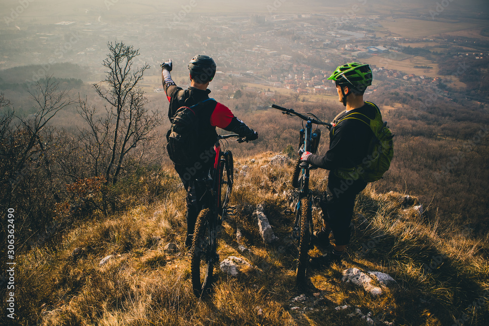 Two men with mountaibikes on a top of the hill. Panorama of a valley with cyclist in the foregroud. One is pointing his fingeer. Two men with mountaibikes on a top of the hill. Panorama of a valley with cyclist in the foregroud. One is pointing his fingeer.