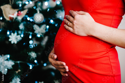 Closeup photo of pregnant woman posing near Christmas tree at home. Merry Christmas and Happy Holidays! Pregnancy, holidays, people and expectation concept.
