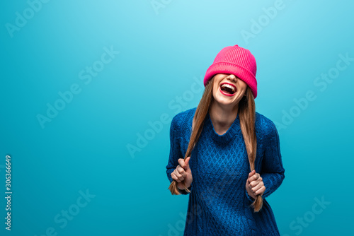 Fotografie, Obraz funny laughing woman in knitted sweater with pink hat on eyes, isolated on blue