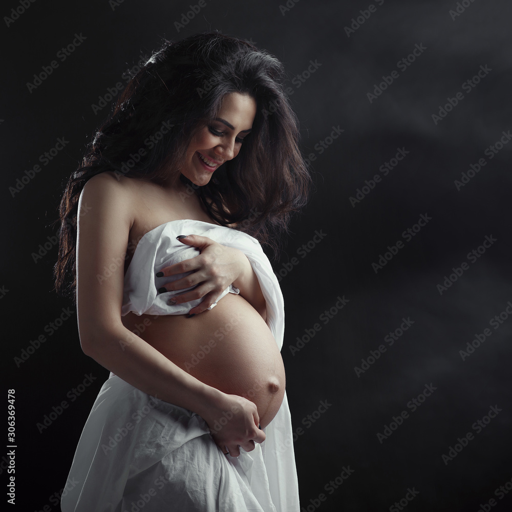 pregnant girl naked in sheet young happy 18 years Caucasian ethnicity  brunette long hair art photo studio dark background beautiful, attractive,  smile, art, strip on the tummy gynecology trimestr Stock Photo |