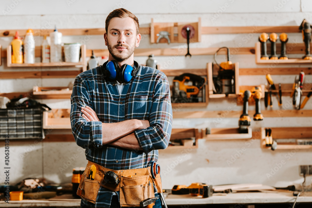 © LIGHTFIELD STUDIOS - handsome carpenter in apron standing with crossed arms in workshop
