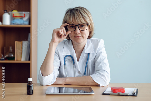 Serious doctor is sitting by her desk and looking ahead.