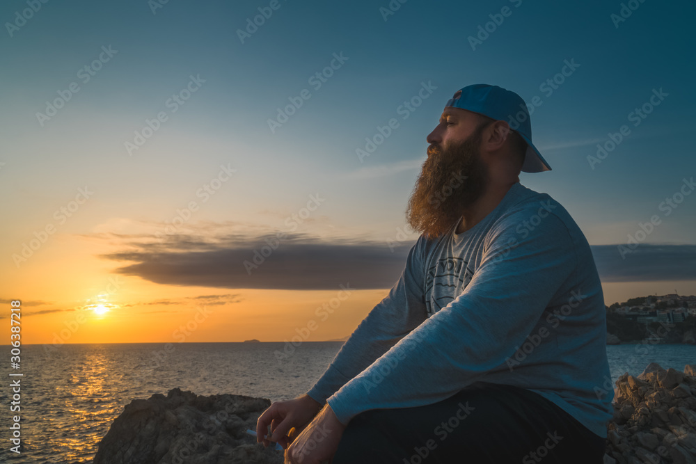 A bearded man is sitting at the rocky shoreline and watching the sea sunset