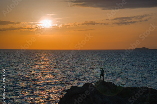 Silhouette of a man staying on the rock and making a photo of sunset into the sea
